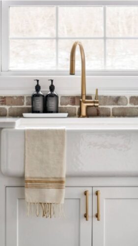 This is a kitchen remodel in east islip new york The was done by lebarron kitchens and photographed by brian phelps. the kitchen has a stunning mahogany range hood. The cabinetry is TW10 doors and crp10 drawers finished in Alpine White. The cabinetry has crown molding and the cabinetry is adorned with gold hardware. The kitchen contains a wine and coffee station with lighting that is easily hidden behind doors. The station has a walnut interior. THe brick wall backsplash adds an organic and timeless feel to the space. The kitchen has a kitchenaid range with a clever narrow pull out spice drawer flanking the side with multitiered drawers next to it. The kitchen has a large porcelain farmhouse sink. The island is home to the kitchenaid microwave drawer, a built in papertowel dispenser for a clutter free countertop and ample seating for four opposite.