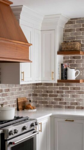 This is a kitchen remodel in east islip new york The was done by lebarron kitchens and photographed by brian phelps. the kitchen has a stunning mahogany range hood. The cabinetry is TW10 doors and crp10 drawers finished in Alpine White. The cabinetry has crown molding and the cabinetry is adorned with gold hardware. The kitchen contains a wine and coffee station with lighting that is easily hidden behind doors. The station has a walnut interior. THe brick wall backsplash adds an organic and timeless feel to the space. The kitchen has a kitchenaid range with a clever narrow pull out spice drawer flanking the side with multitiered drawers next to it. The kitchen has a large porcelain farmhouse sink. The island is home to the kitchenaid microwave drawer, a built in papertowel dispenser for a clutter free countertop and ample seating for four opposite.