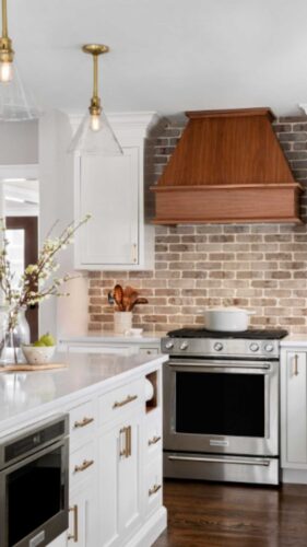 This is a kitchen remodel in east islip new york The was done by lebarron kitchens and photographed by brian phelps. the kitchen has a stunning mahogany range hood. The cabinetry is TW10 doors and crp10 drawers finished in Alpine White. The cabinetry has crown molding and the cabinetry is adorned with gold hardware. The kitchen contains a wine and coffee station with lighting that is easily hidden behind doors. The station has a walnut interior. THe brick wall backsplash adds an organic and timeless feel to the space. The kitchen has a kitchenaid range with a clever narrow pull out spice drawer flanking the side with multitiered drawers next to it. The kitchen has a large porcelain farmhouse sink. The island is home to the kitchenaid microwave drawer, a built in papertowel dispenser for a clutter free countertop and ample seating for four opposite.