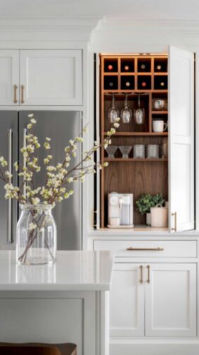 This is a kitchen remodel in east islip new york The was done by lebarron kitchens and photographed by brian phelps. the kitchen has a stunning mahogany range hood. The cabinetry is TW10 doors and crp10 drawers finished in Alpine White. The cabinetry has crown molding and the cabinetry is adorned with gold hardware. The kitchen contains a wine and coffee station with lighting that is easily hidden behind doors. The station has a walnut interior. THe brick wall backsplash adds an organic and timeless feel to the space. The kitchen has a kitchenaid range with a clever narrow pull out spice drawer flanking the side with multitiered drawers next to it. The kitchen has a large porcelain farmhouse sink. The island is home to the kitchenaid microwave drawer, a built in papertowel dispenser for a clutter free countertop and ample seating for four opposite.