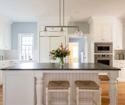A white kitchen with pale blue walls, a kitchen island with seating and wolf wall ovens