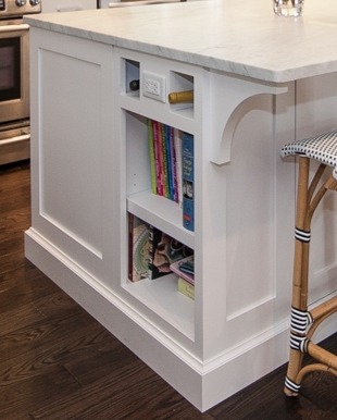 This is a picture of a small white bookcase made under the island for a white kitchen featuring custom made cabinetry by Kountry Kraft Cabinetry. 