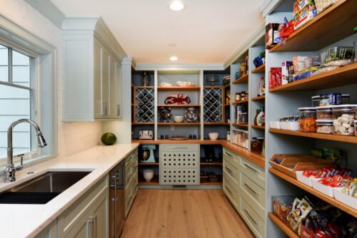Walk in pantry with soft green custom kitchen cabinets, open wood shelves stocked with snacks and jars, wine rack cubbies, and a sink with white countertop and light wood floor.