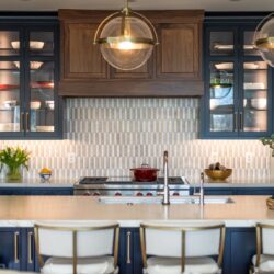 Kitchen remodel in Annapolis Maryland featuring custom cabinetry, walnut accents, and navy finishes by Kountry Kraft and Chroma Kitchens. Walnut range hood framed by navy finished upper cabinetry with glass doors. Navy finished island with seating for four. White countertops.