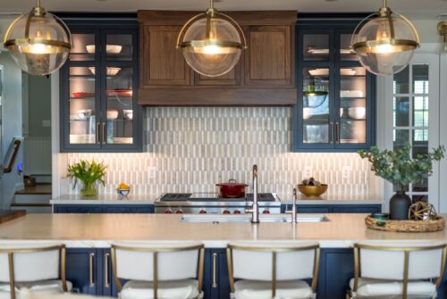 Kitchen remodel in Annapolis Maryland featuring custom cabinetry, walnut accents, and navy finishes by Kountry Kraft and Chroma Kitchens. Walnut range hood framed by navy finished upper cabinetry with glass doors. Navy finished island with seating for four. White countertops.