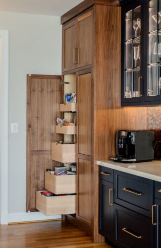 Kitchen remodel in Annapolis Maryland featuring custom cabinetry, walnut accents, and navy finishes by Kountry Kraft and Chroma Kitchens. Walnut range hood framed by navy finished upper cabinetry with glass doors. Navy finished island with seating for four. White countertops. Dry bar area showing tall pantry cabinet with pull out drawers.
