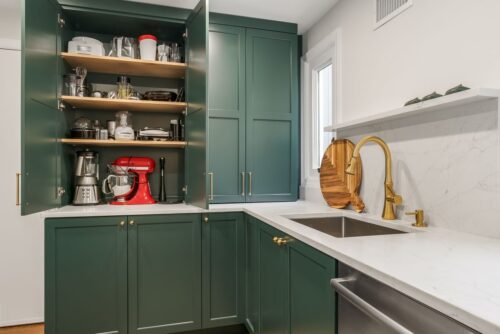 Custom kitchen Short Hills NJ picture showing butler's pantry with green painted cabinetry and honey bronze fixtures. There is a sink and dishwasher. Upper cabinet doors are open showing storage.