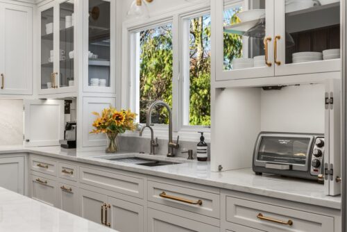 Custom kitchen Short Hills NJ with white perimeter cabinetry. Picture is showing sink area with honey bronze cabinet pulls and glass paneled doors on the upper cabinets