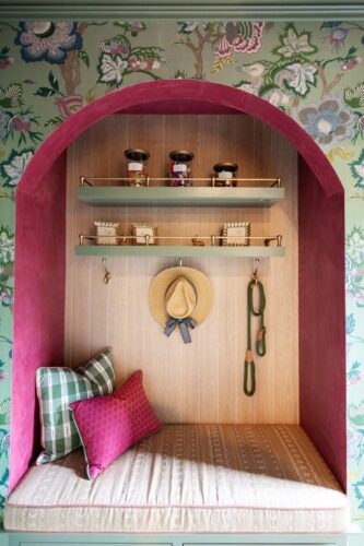 Bench nook with green cabinetry, straw hat, pillows, and floral wallpaper in the Designer Show House mudroom.