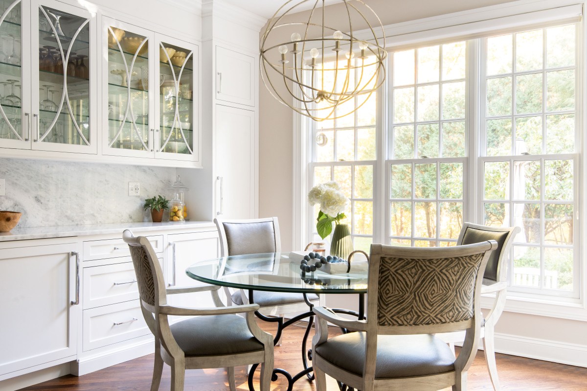 White custom dining room built-ins featuring glass-front display cabinets with lighting, a marble countertop, and an integrated dining area with a glass table and geometric chairs.