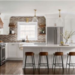 This is a kitchen remodel in east islip new york The was done by lebarron kitchens and photographed by brian phelps. the kitchen has a stunning mahogany range hood. The cabinetry is TW10 doors and crp10 drawers finished in Alpine White. The cabinetry has crown molding and the cabinetry is adorned with gold hardware. The kitchen contains a wine and coffee station with lighting that is easily hidden behind doors. The station has a walnut interior. THe brick wall backsplash adds an organic and timeless feel to the space. The kitchen has a kitchenaid range with a clever narrow pull out spice drawer flanking the side with multitiered drawers next to it. The kitchen has a large porcelain farmhouse sink. The island is home to the kitchenaid microwave drawer, a built in papertowel dispenser for a clutter free countertop and ample seating for four opposite.