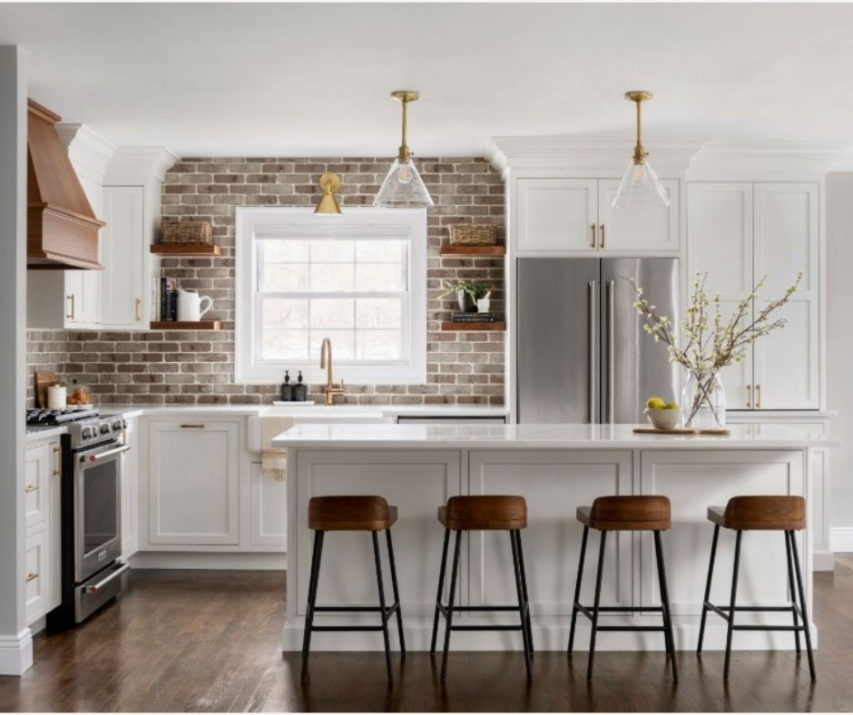 This is a kitchen remodel in east islip new york The was done by lebarron kitchens and photographed by brian phelps. the kitchen has a stunning mahogany range hood. The cabinetry is TW10 doors and crp10 drawers finished in Alpine White. The cabinetry has crown molding and the cabinetry is adorned with gold hardware. The kitchen contains a wine and coffee station with lighting that is easily hidden behind doors. The station has a walnut interior. THe brick wall backsplash adds an organic and timeless feel to the space. The kitchen has a kitchenaid range with a clever narrow pull out spice drawer flanking the side with multitiered drawers next to it. The kitchen has a large porcelain farmhouse sink. The island is home to the kitchenaid microwave drawer, a built in papertowel dispenser for a clutter free countertop and ample seating for four opposite.