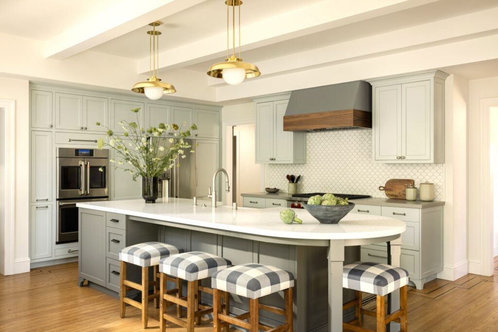 Floor to ceiling kitchen cabinets in a soft sage green finish paired with a large white island, brass pendant lighting, and a custom wood-accent range hood in a bright, elegant kitchen design
