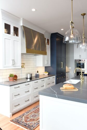 White and blue kitchen cabinets with a custom brushed stainless steel range hood with matte brass straps and rivets — a focal point that blends traditional and contemporary styles seamlessly. 