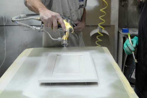 Craftsman applying a professional sprayed finish to a custom cabinet door in the Kountry Kraft finishing shop, highlighting precision and quality.