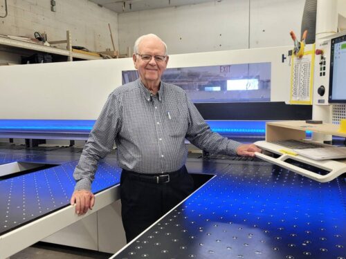Elvin Hurst Sr. standing in the Kountry Kraft cabinet makers workshop beside modern woodworking equipment.