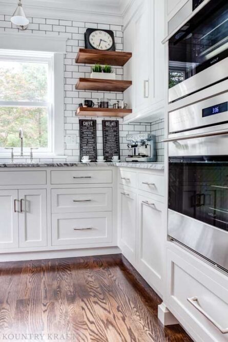 Custom inset white perimeter cabinets with subway tile, marble countertops floating shelves and stainless steel appliances