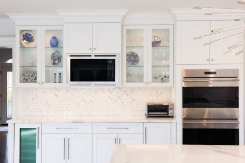 Glass-front display cabinets and built-in microwave with herringbone backsplash in Brookville NY kitchen remodel.